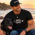 Hombre sonriendo con su perro negro, vistiendo una camiseta negra Dog Dad en la playa al atardecer.